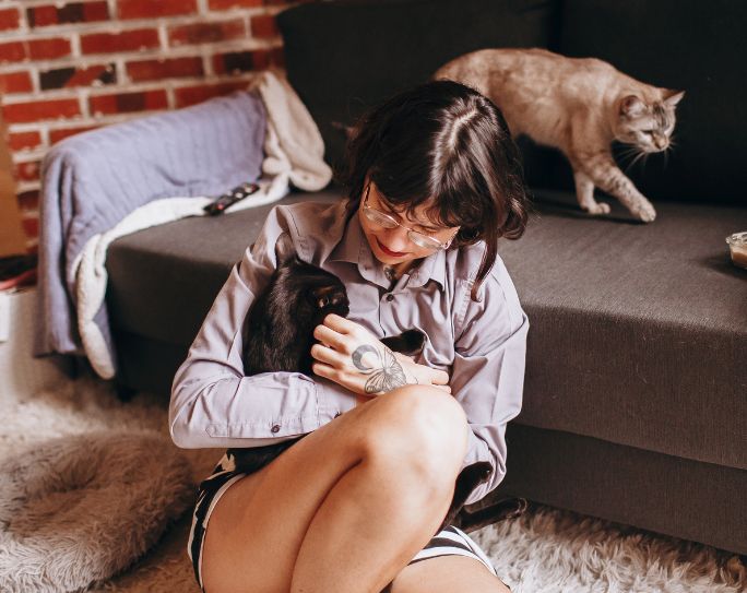Woman pet sitter sitting on floor in front of couch holding cat, while her other cat walks across the couch behind her.